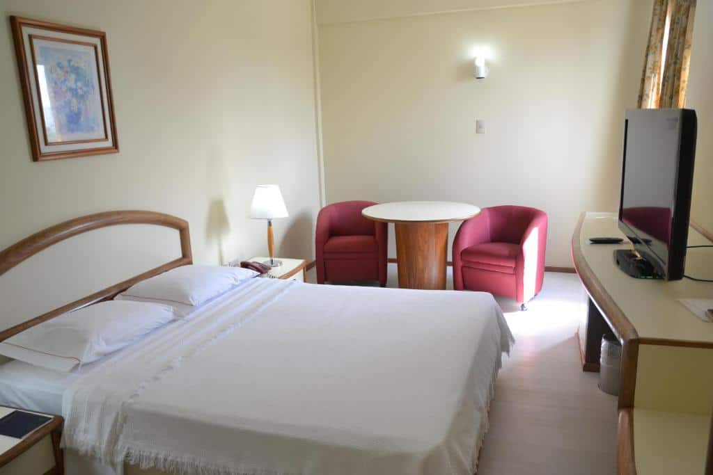 Quarto no Hotel Sagres com uma cama de casal, duas cadeiras vermelhas ao redor de uma mesa redonda, uma TV e uma luminária de cabeceira, para representar hotéis em Belém do Pará.