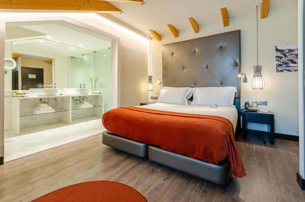 Quarto do Hotel Santa Justa com uma cama de casal, cobertor laranja, área de banheiro aberta com pias duplas e piso de madeira, para representar hotéis no Chiado em Lisboa.