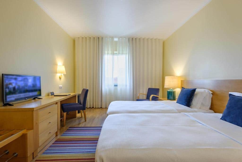 Quarto do Hotel Santa Maria com duas camas de solteiro, uma escrivaninha com cadeira, TV, poltrona, abajur e janela com cortinas brancas, para representar onde ficar em Fátima.