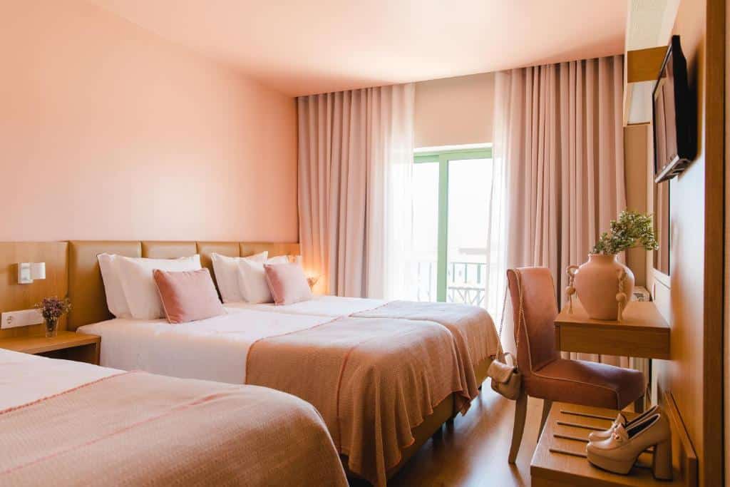 Quarto no Hotel Santo Antônio de Fátima com duas camas de casal, paredes cor-de-rosa, roupa de cama combinando, uma escrivaninha com um vaso e luz do sol entrando pela janela, para representar onde ficar em Fátima.