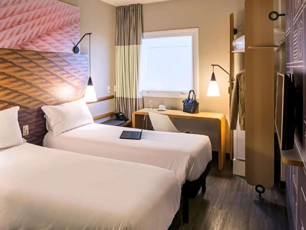 Um quarto no ibis São Paulo Tatuape com duas camas de solteiro, uma escrivaninha de parede, um laptop, um cabide e uma janela com cortinas fechadas, para representar hotéis ibis em São Paulo.