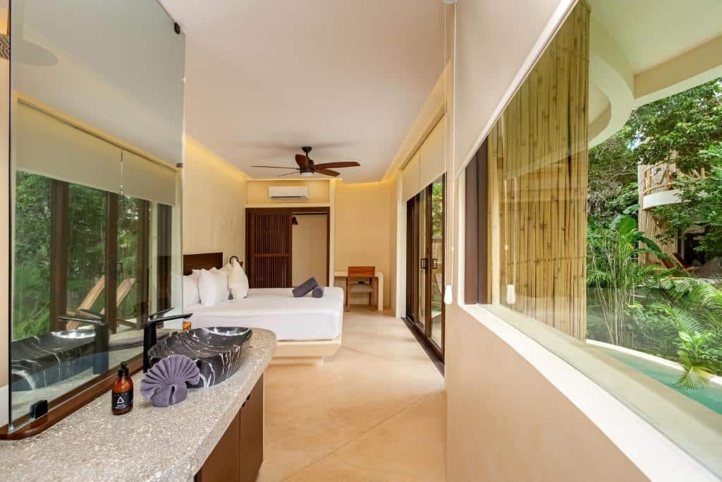 Quarto moderno no Kan Tulum com cama, escrivaninha, ventilador de teto, janelas grandes e vista para uma piscina privativa e vegetação externa, para representar hotéis em Tulum.