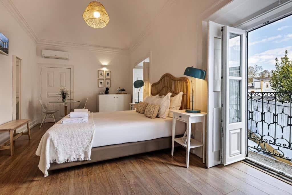 Quarto bem iluminado no Lavradores Boutique Guesthouse com cama de casal, roupa de cama branca, mesas de cabeceira e portas de varanda abertas com vista para árvores e edifícios, para representar onde ficar em Évora.