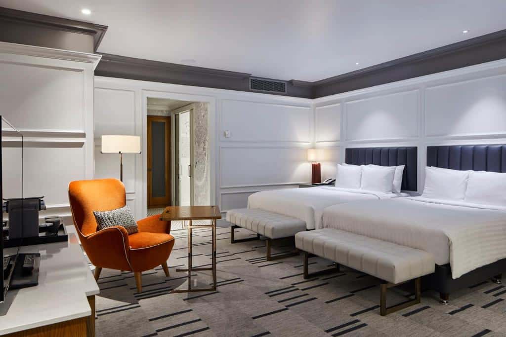 Quarto no London Marriott Hotel County Hall com duas camas grandes, uma cadeira laranja, uma escrivaninha, uma TV e uma decoração minimalista em cores neutras.