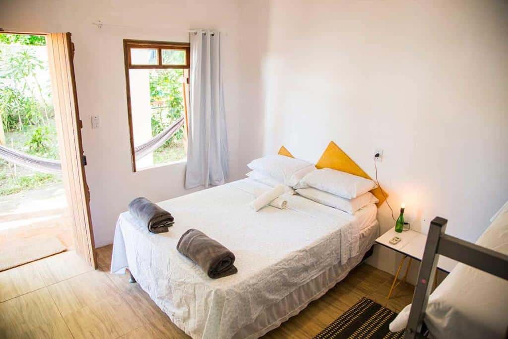 Um quarto arrumado com uma cama de casal, toalhas dobradas, uma pequena mesa de cabeceira, uma janela e uma rede visível do lado de fora, no Mangue Seco Suítes & Bistrô em Mangue Seco, para representar pousadas na Linha Verde na Bahia.