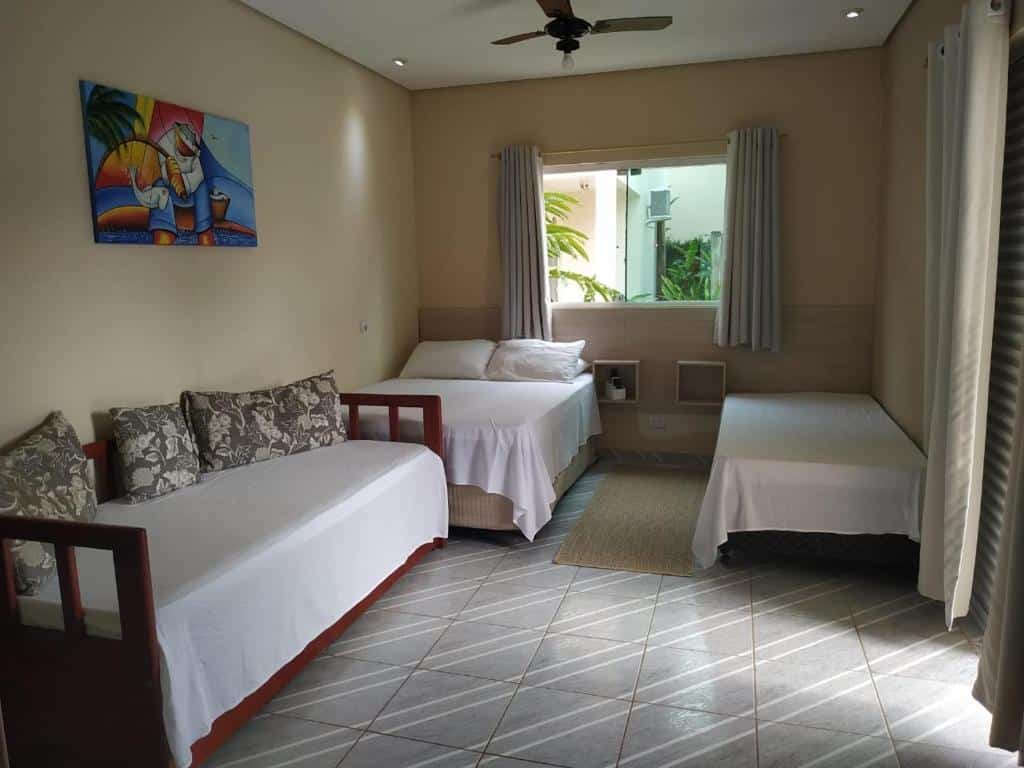 Quarto no Mares de Camburizinho com uma cama de casal, duas camas de solteiro, um ventilador de teto, obras de arte na parede e uma janela com cortinas, para representar airbnb na Praia da Baleia.