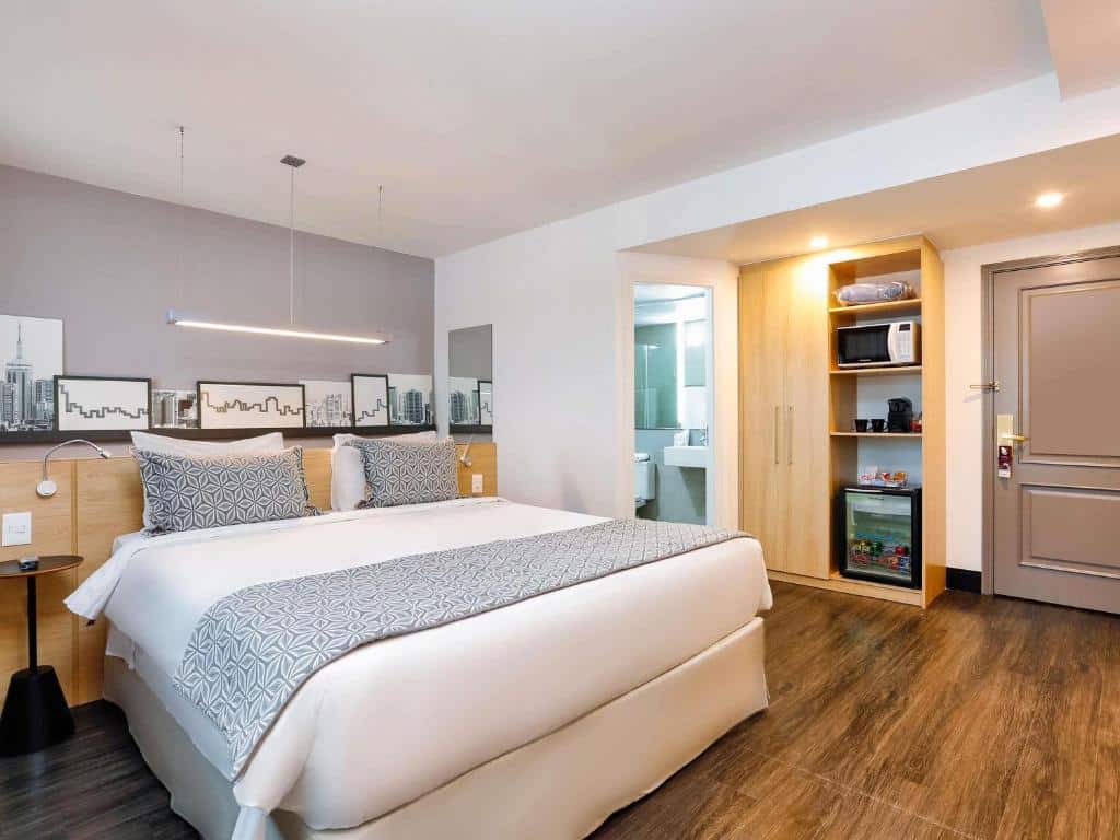 Quarto moderno no Mercure São Paulo Pinheiros com uma cama de casal, arte na parede, armário aberto, frigobar, micro-ondas e vista para o banheiro, para representar hotéis perto do Allianz Parque.