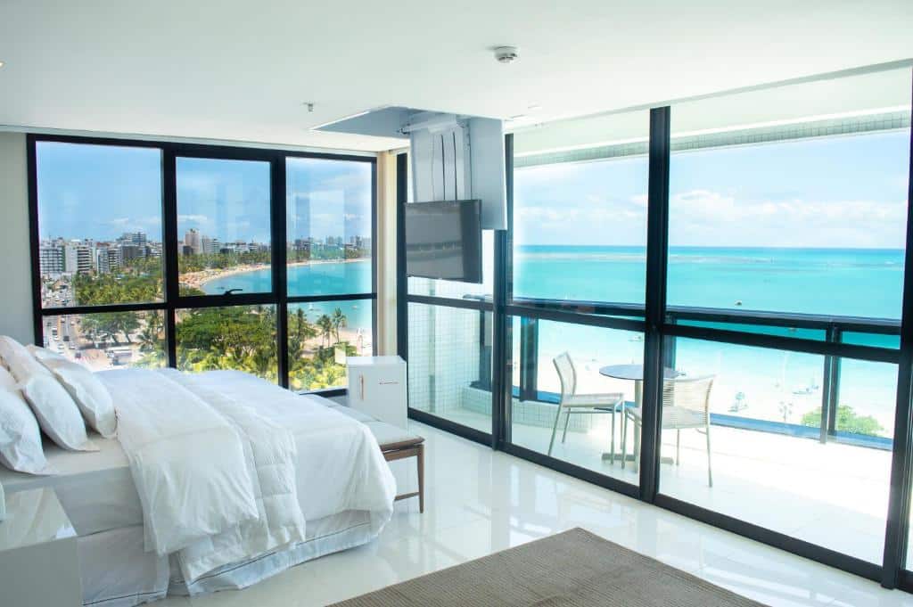 Quarto moderno no Meridiano Hotel em Maceió, com roupa de cama branca, janelas grandes, vista para o mar, varanda e uma pequena mesa externa com duas cadeiras.