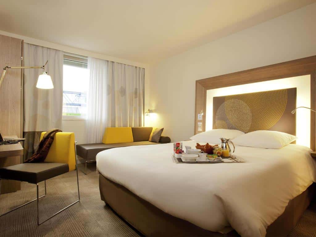 Quarto no Novotel Paris Gare De Lyon com uma cama de casal, escrivaninha, cadeira, sofá e uma bandeja de café da manhã sobre a cama, perto de uma janela com cortinas, para representar hotéis com piscina em Paris.