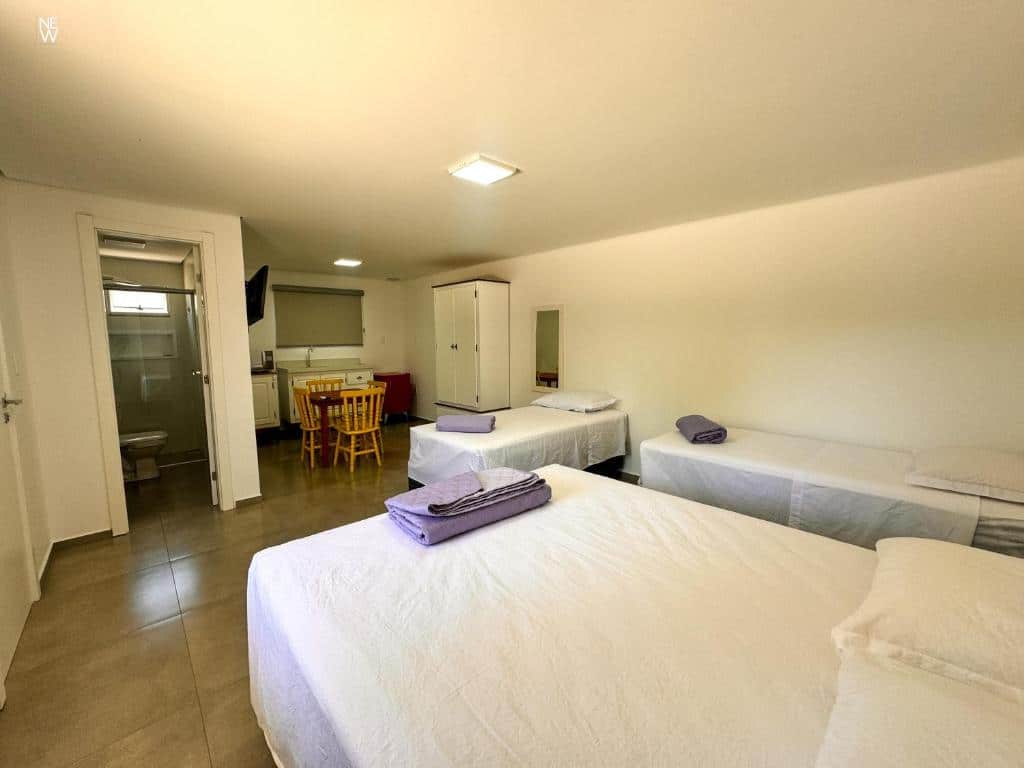 Um quarto bem iluminado com quatro camas, uma pequena mesa de jantar, um guarda-roupa, uma TV e uma porta aberta para um banheiro, no OPY CAMBOA, para representar pousadas em Governador Celso Ramos.