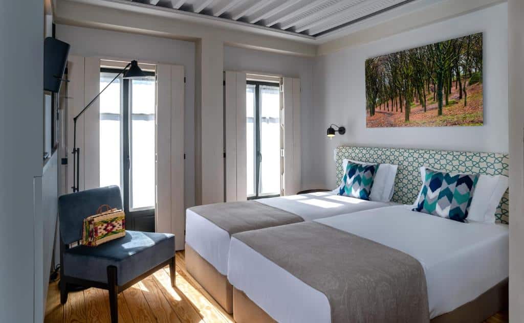 Um quarto no Porta Nova Collection House bem iluminado com duas camas de solteiro, uma cadeira azul, arte na parede e janelas grandes que permitem a entrada de luz natural, para representar hotéis em Braga.
