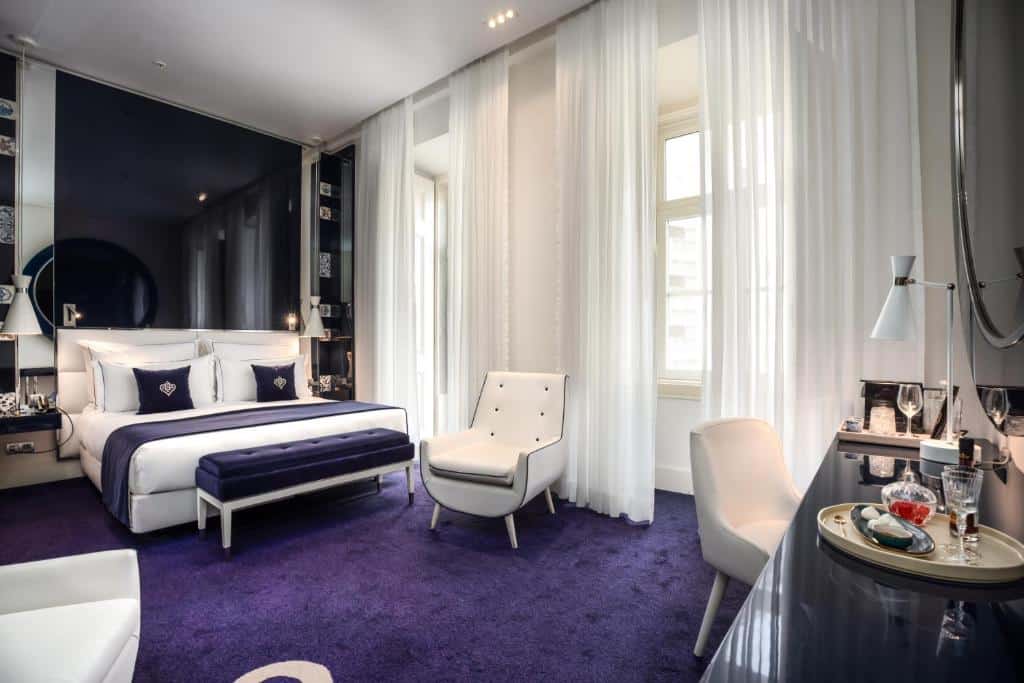 Quarto no Portugal Boutique Hotel com carpete roxo, cama branca, poltrona, escrivaninha e janelas grandes com cortinas brancas transparentes, paa representar hotéis no Chiaod em Lisboa.