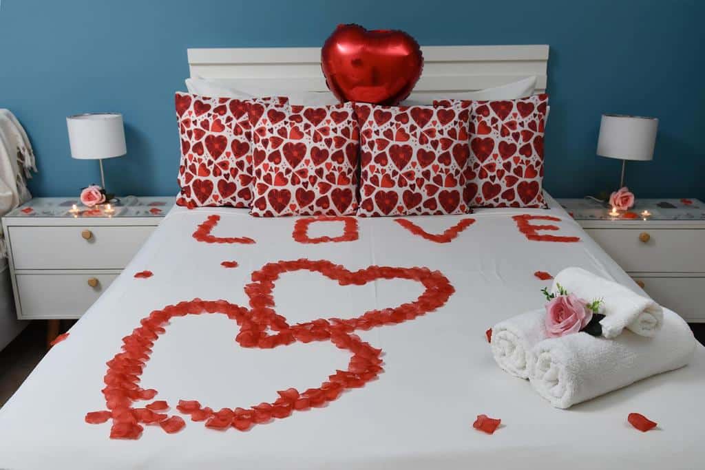Uma cama decorada com pétalas de rosas vermelhas em forma de coração e a palavra "LOVE" (amor), travesseiros com estampas de coração e um balão de coração, na Pousada Rosa dos Ventos Juquehy, para representar pousadas em Juquehy.