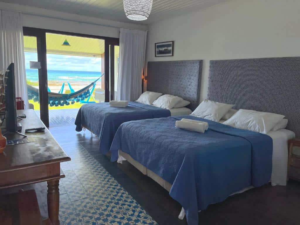 Um quarto na Pousada a Capela em Arembepe, com duas camas, roupa de cama azul, uma escrivaninha e uma rede em um pátio de frente para o oceano, para representar pousadas na Linha Verde na Bahia.