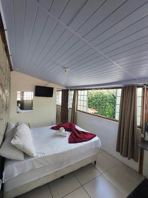 Um pequeno quarto na Pousada Acayaca com uma cama de casal, roupa de cama branca, cobertor vermelho, TV, espelho e janelas com cortinas marrons.