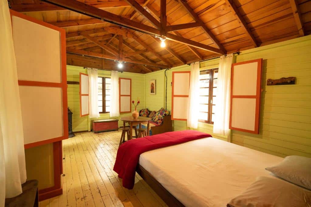 Um quarto de madeira aconchegante com uma cama de casal, uma mesa pequena e janelas com cortinas brancas e detalhes em laranja, iluminadas por luzes no teto, na Pousada Aguaraguazu em Tibagi, para representar pousadas no interior do Paraná.
