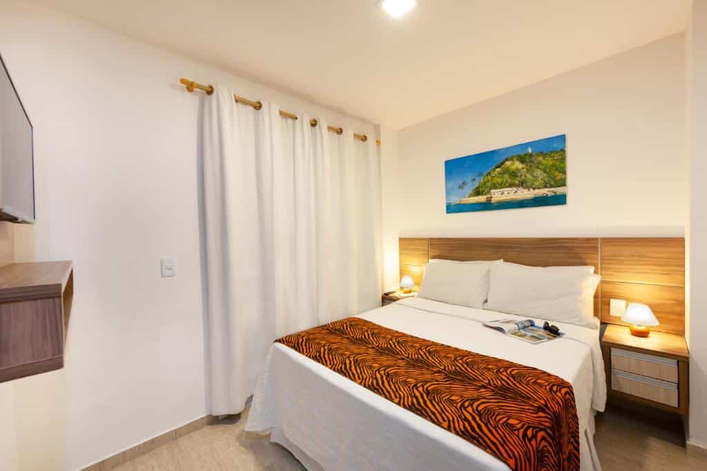 Uma cama bem arrumada com um cobertor laranja estampado em um quarto pequeno e bem iluminado com paredes brancas e uma foto de praia na parede, na Pousada Bahia 10, para representar pousadas em Morro de São Paulo.