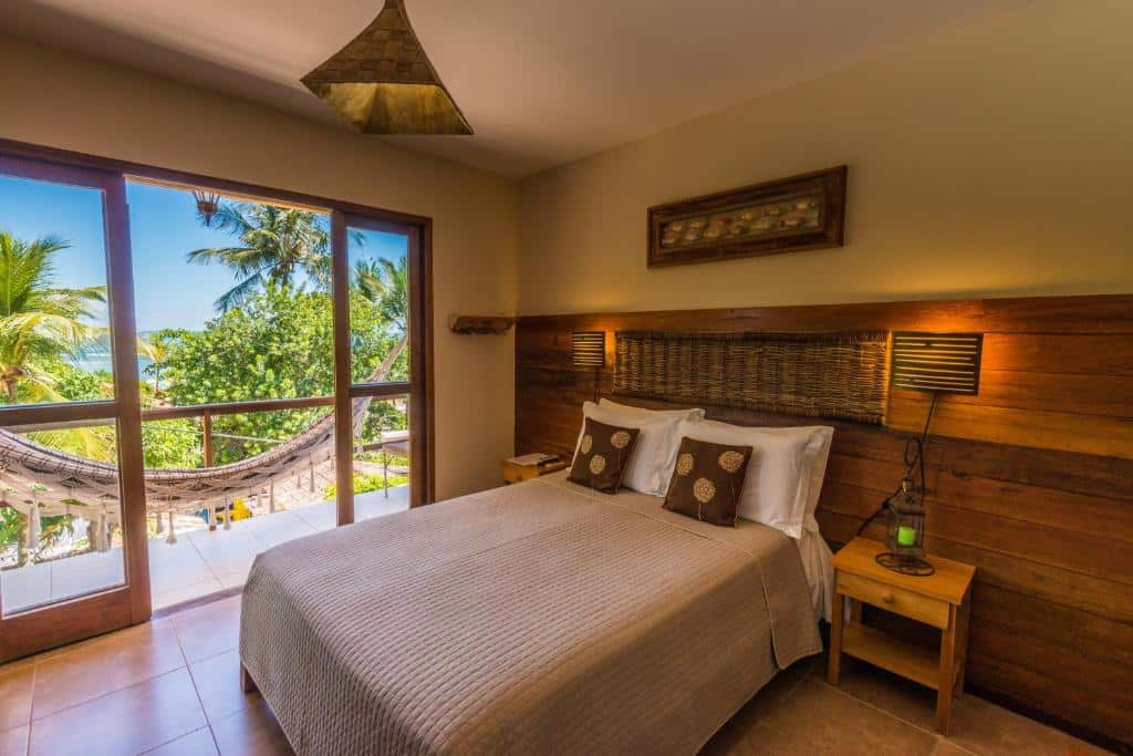 Um quarto aconchegante com uma cama de casal, detalhes em madeira, mesas de cabeceira e uma varanda com rede e vista tropical, na Pousada Bahia Bella, para representar pousadas em Morro de São Paulo.