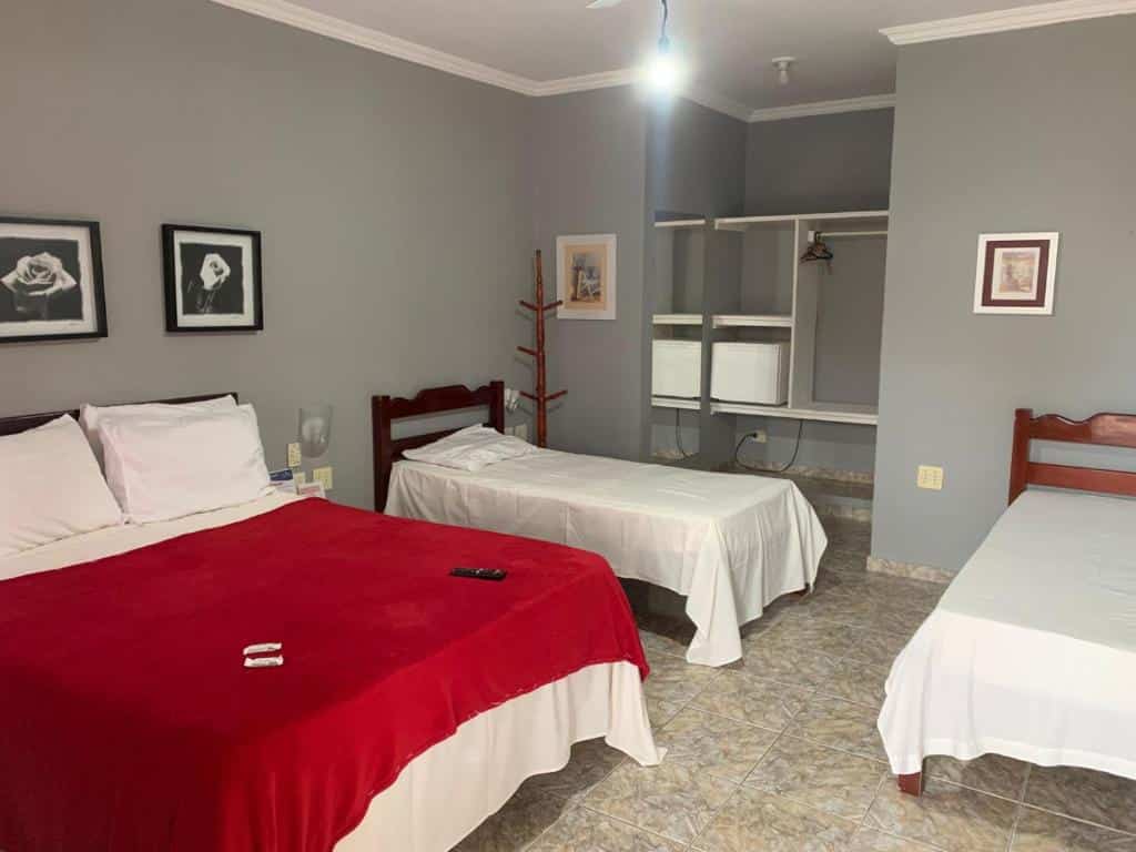 Um quarto na Pousada Belmare Eireli com uma cama de casal, uma cama de solteiro, um controle remoto de TV, espaço aberto no armário e paredes cinza com arte emoldurada, para representar pousadas em Rio das Ostras.