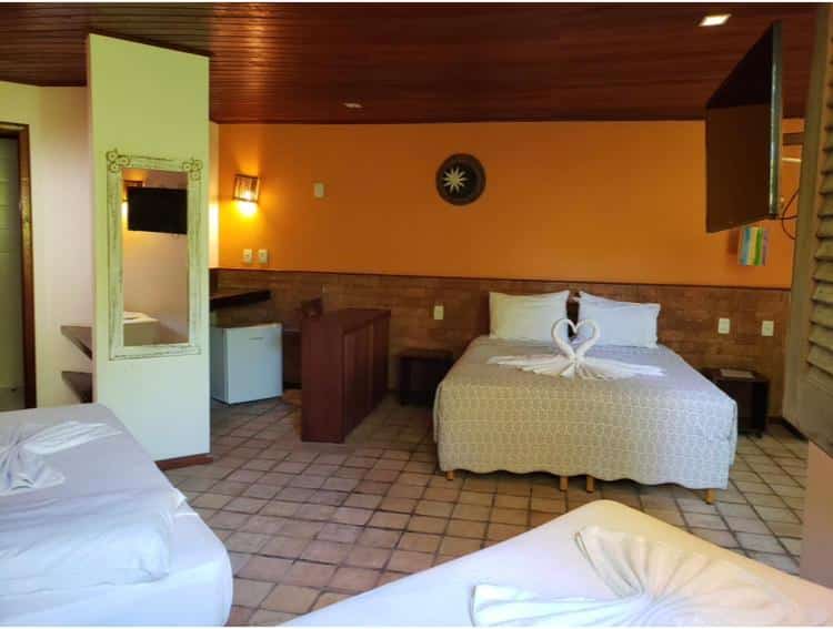 Quarto na Pousada Bichelenga com duas camas, um frigobar pequeno, um espelho, uma TV e cisnes de toalha nas camas, com decoração laranja e marrom, para representar pousadas em Imbassaí.