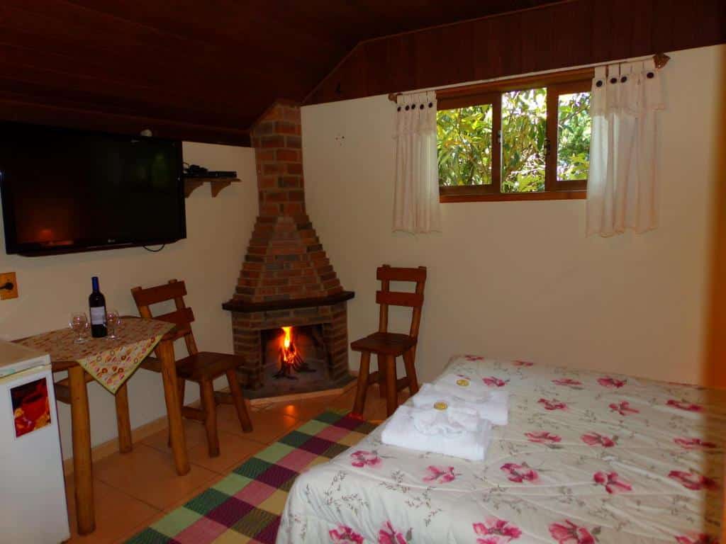 Quarto pequeno e aconchegante na Pousada Caminho do Escorrega com uma cama, lareira de tijolos, TV, duas cadeiras de madeira, frigobar, janela e uma mesa com vinho e taças, para representar pousadas em Visconde de Mauá.
