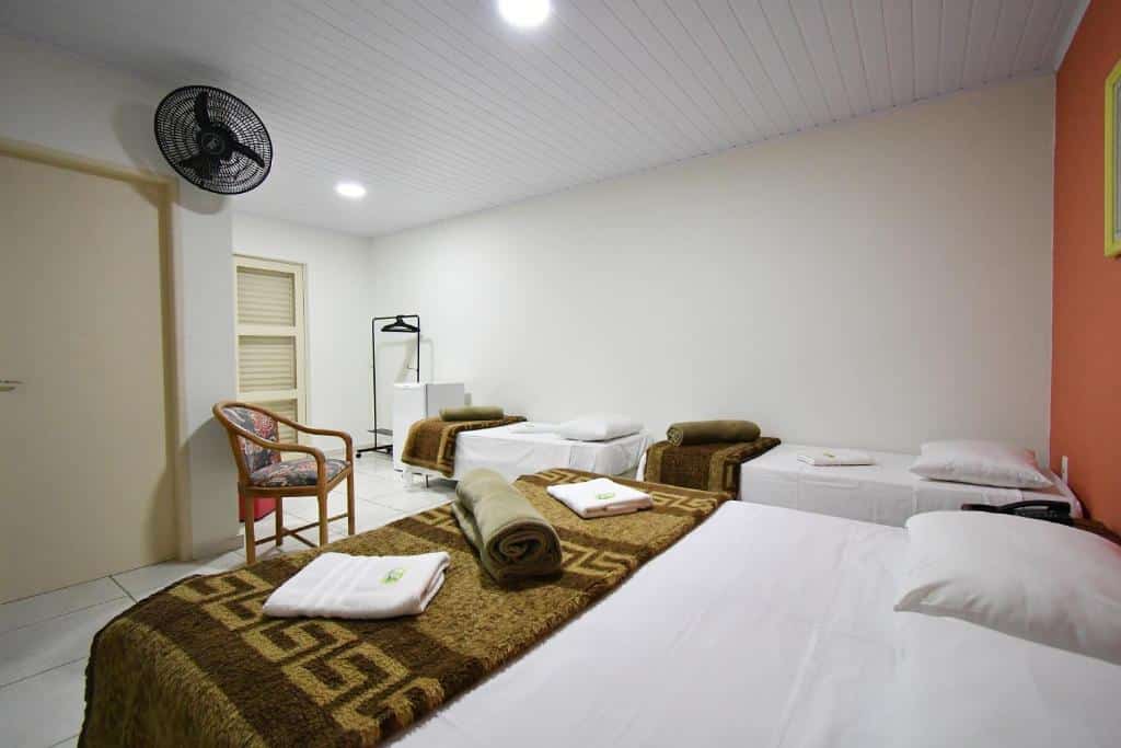 Um quarto da Pousada Casa Doce Vida com uma cama de casal, duas camas de solteiro, toalhas dobradas, uma cadeira, um ventilador e um cabideiro, para representar pousadas em Águas de Lindóia.