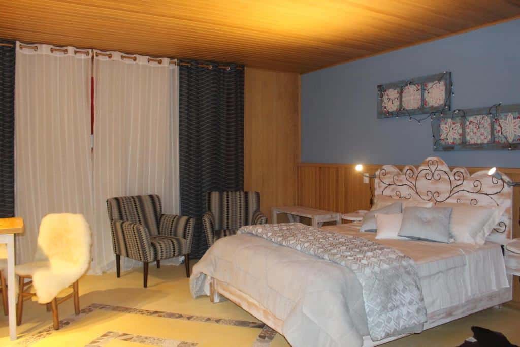 Um quarto aconchegante na Pousada dos Cataventos, com uma cama de casal, poltronas listradas, uma escrivaninha com uma cadeira coberta de pele e cortinas estampadas, para representar pousadas em São Francisco de Paula.