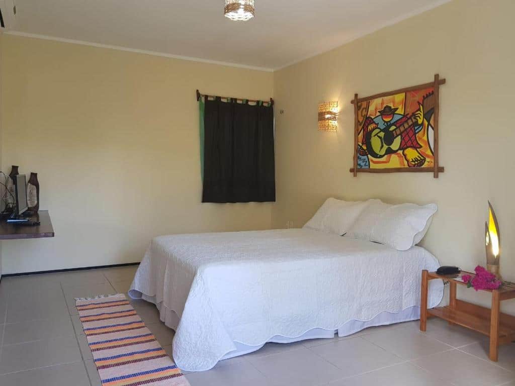 Um quarto simples com uma cama branca, arte de parede colorida, uma pequena mesa de cabeceira e um tapete listrado em um piso de azulejos, na Pousada Coqueiro Beach, para representar pousadas em Luís Correia.