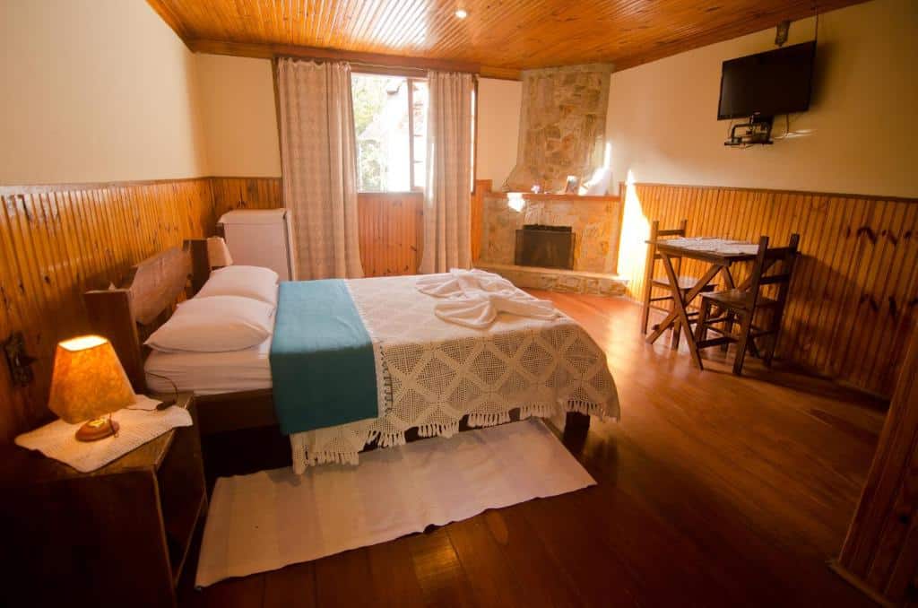 Quarto na Pousada da Carmem com painéis de madeira com uma cama de casal, uma pequena mesa e cadeiras, uma lareira, TV, abajur e janela com cortinas, para representar pousadas em Visconde de Mauá.