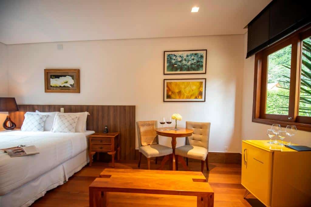 Um quarto aconchegante na Pousada Déllis com uma cama, uma mesa de madeira com duas cadeiras, arte na parede, uma janela e um pequeno frigobar amarelo, para representar pousadas em Visconde de Mauá.