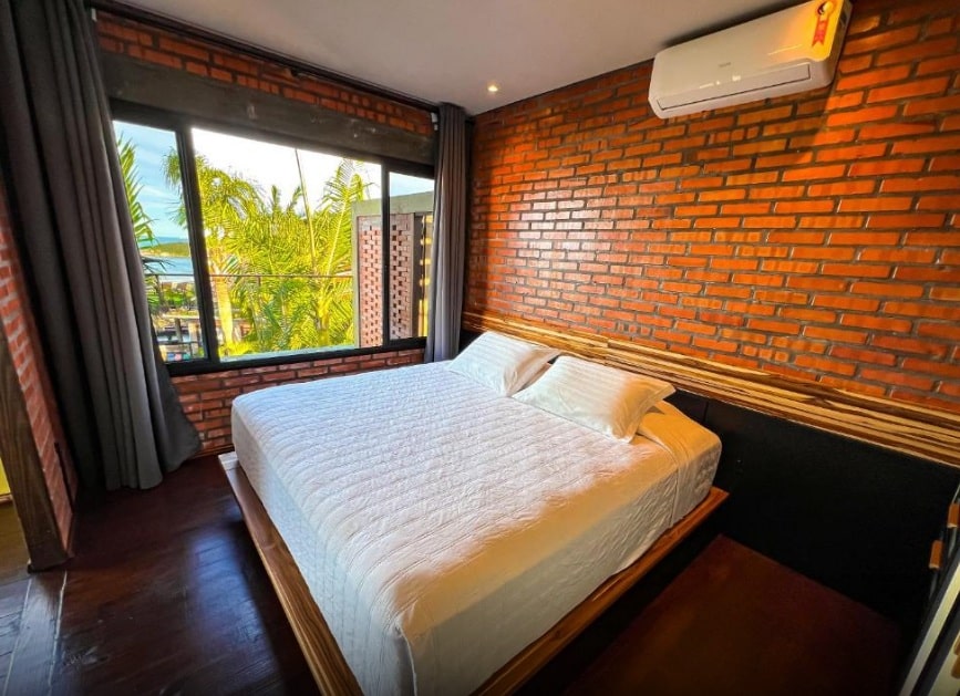 Um quarto com uma parede de tijolos, uma cama grande com um edredom branco e uma janela com vegetação externa, na Pousada Descanso do Rei, para representar pousadas na Praia do Rosa.