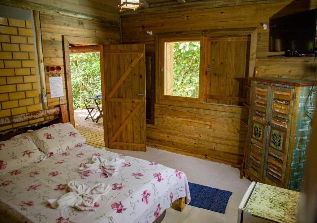 Um quarto rústico de madeira com colcha floral, porta e janela abertas, armário, TV e vista para a vegetação externa, na Pousada Favo de Mel na Ilha do Mel, para representar pousadas no interior do Paraná.