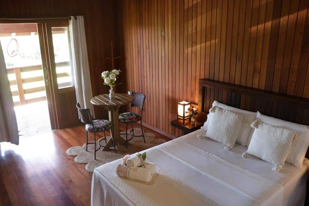 Um quarto de madeira na Pousada Gauleses com uma cama bem arrumada, uma pequena mesa redonda com duas cadeiras e uma porta aberta que dá para uma varanda, para representar pousadas na Praia do Rosa.