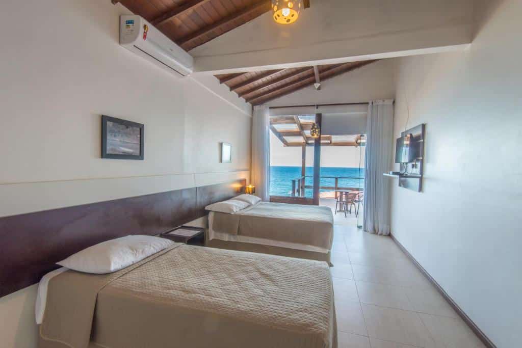 Quarto na Pousada Ilha da Saudade com duas camas, TV e portas de pátio que se abrem para uma varanda com vista para o mar sob um teto de madeira, para representar pousadas em Morro de São Paulo.