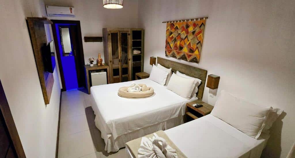 Um quarto na Pousada João Sol na Praia do Forte, com uma cama de casal e uma cama de solteiro, móveis de madeira, frigobar e um enfeite de parede estampado, para representar pousadas na Linha Verde na Bahia.