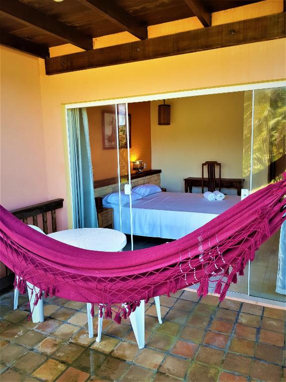Uma rede vermelha está pendurada em um pátio de azulejos do lado de fora de um quarto com cama, mesa e cadeira, visto através de portas de vidro deslizantes, na Pousada Luar da Praia, para representar pousadas em Imbassaí.