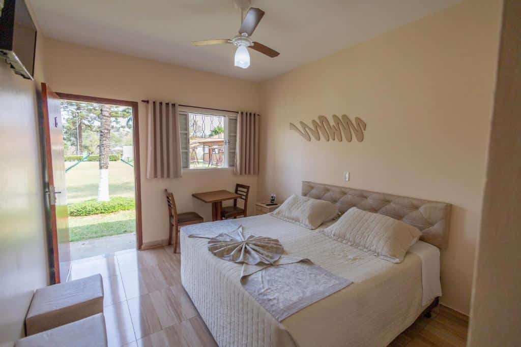 Um quarto bege na Pousada Mirante das Estancias com uma cama de casal, mesa pequena com duas cadeiras, janela, porta aberta para um pátio gramado e ventilador de teto, para representar pousadas em Águas de Lindóia.