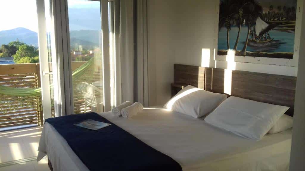 Quarto iluminado pelo sol na Pousada Morro do Rosa com uma cama de casal, toalhas enroladas, varanda, rede e uma pintura na parede acima da cabeceira da cama, para representar pousadas na Praia do Rosa.