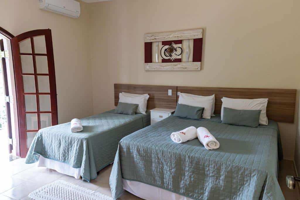 Um quarto no Pousada Nefelibatas com duas camas, colchas verdes, toalhas enroladas em cada uma e uma obra de arte emoldurada acima das cabeceiras, para representar pousadas em Águas de Lindóia.