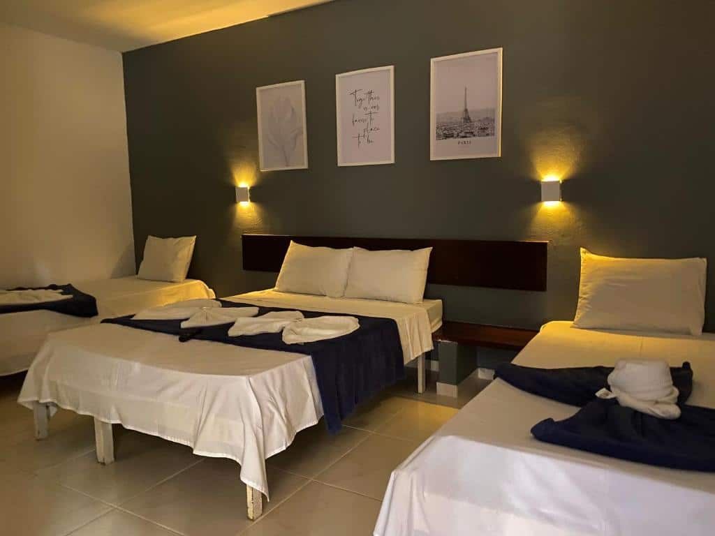 Três camas de solteiro na Pousada Puerta Del Sol Rio das Ostras, bem arrumadas com lençóis brancos e cobertores azul-marinho em um quarto pouco iluminado com arte na parede acima da cabeceira, para representar pousadas em Rio das Ostras.