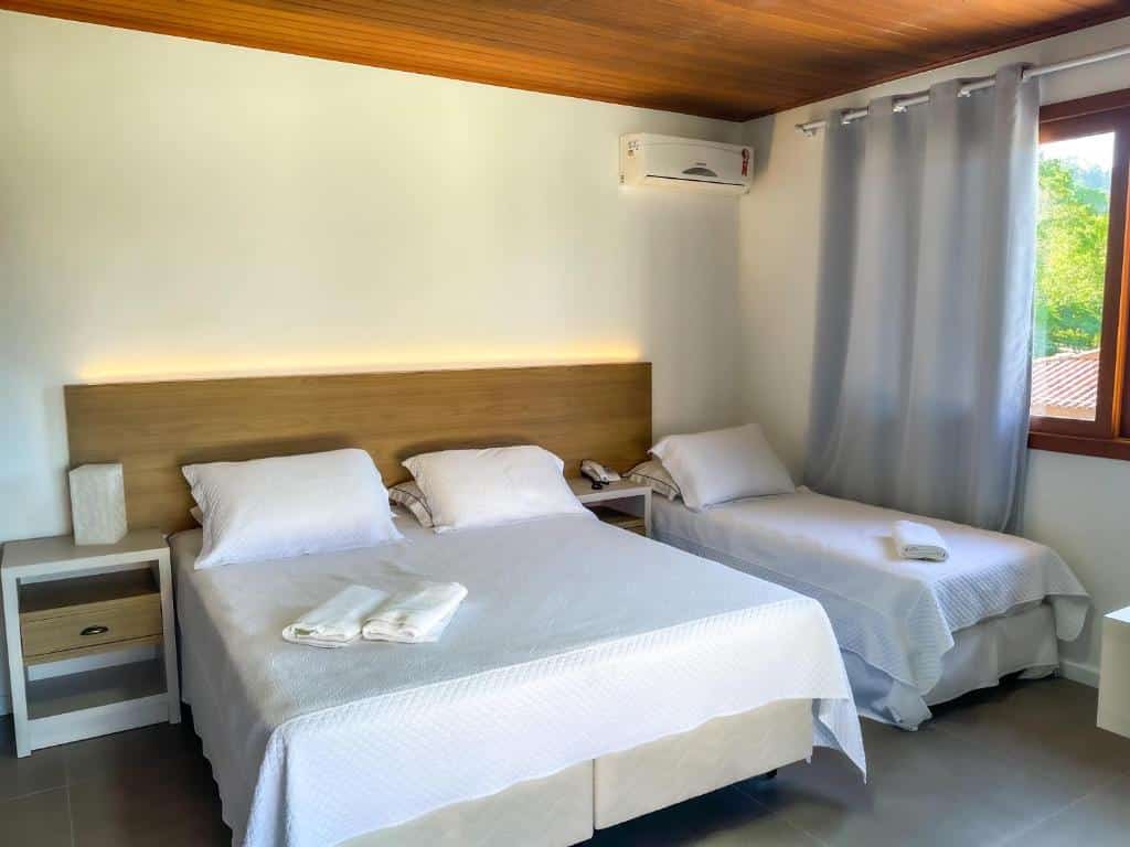 Quarto na Pousada Recanto das Flores com uma cama de casal e uma cama de solteiro, ambas com roupa de cama branca, mesinhas de cabeceira e uma grande janela com cortinas, para representar pousadas em Governador Celso Ramos.