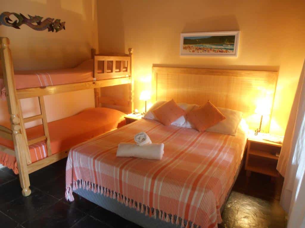Um quarto com uma cama de casal e um beliche, ambos com roupa de cama laranja, mesas de cabeceira e arte na parede acima da cama, na Pousada Rosa & Canela, para representar pousadas na Praia do Rosa.