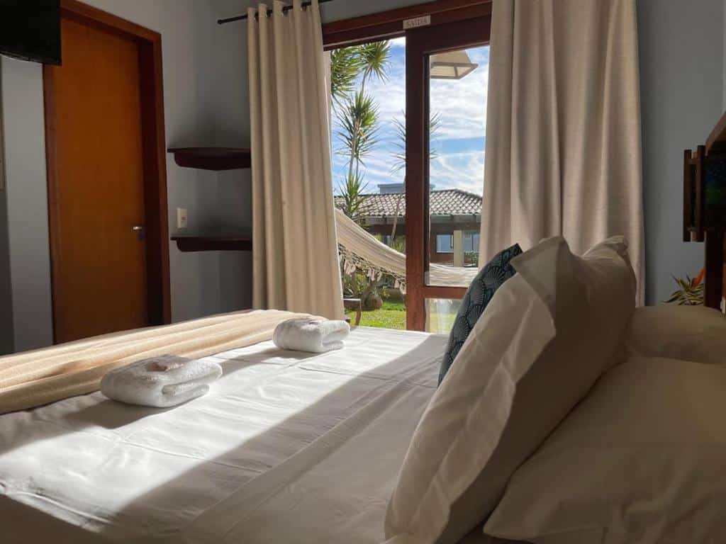 Uma cama bem arrumada com toalhas fica de frente para uma janela com cortinas, mostrando uma rede e um jardim externo à luz do dia, no Rosamar Suites, para representar pousadas na Praia do Rosa.