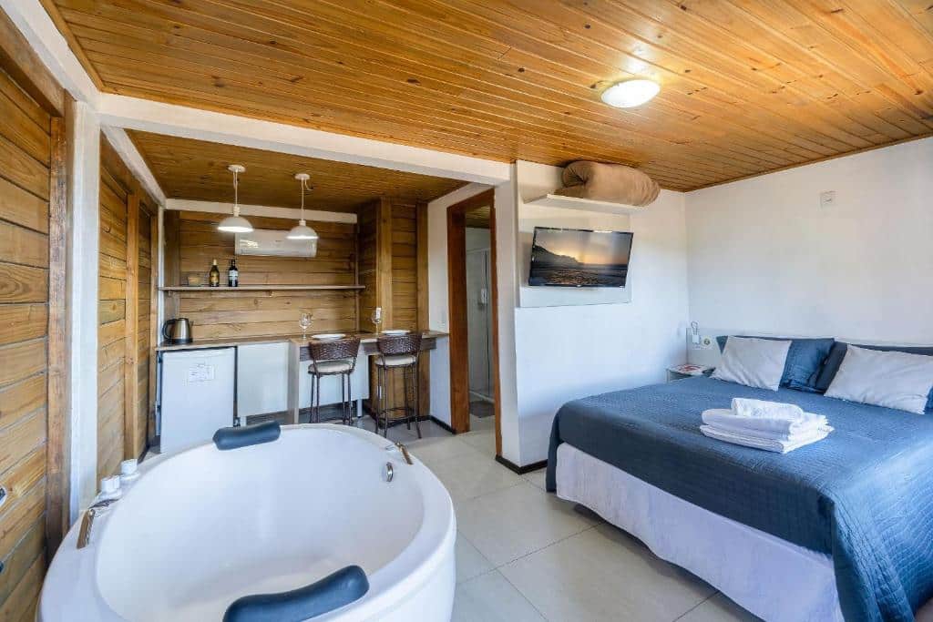 Um quarto na Pousada Solar dos Lírios com uma cama, TV, banheira redonda, área de bar, duas banquetas, painéis de madeira e toalhas dobradas sobre a cama, para representar pousadas na Praia do Rosa.