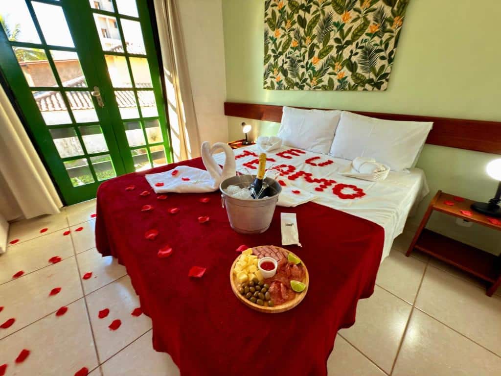 Um quarto na Pousada Sonho Verde com cama de casal, cobertor vermelho, pétalas de rosa, cisnes de toalha, bandeja de frutas e vinho em um balde de gelo, paa representar pousadas no Rio das Ostras.