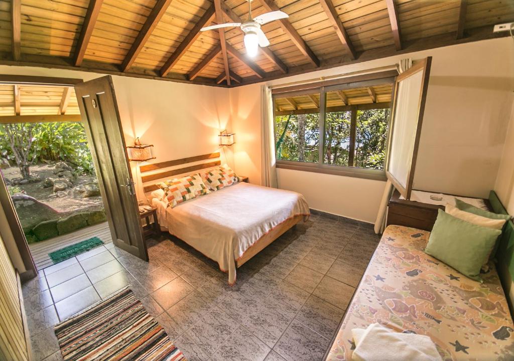 Um quarto na Pousada The Rosebud com uma cama de casal, uma espreguiçadeira, piso de azulejos, teto de madeira, ventilador de teto e janelas grandes com vista para o jardim, para representar pousadas na Praia do Rosa.