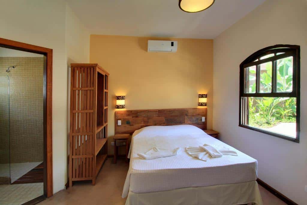 Um quarto simples na Pousada Tupinamba com uma cama de casal, móveis de madeira, luminárias de parede e uma janela com vegetação externa. Banheiro à esquerda, para representar pousadas em Juquehy.