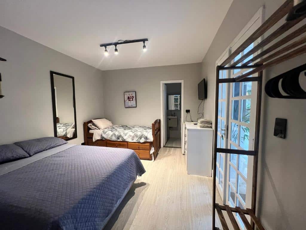 Um quarto na Pousada Vale Beach com uma cama de casal, uma cama de solteiro, um espelho de parede, TV, cômoda, armário aberto e portas francesas, para representar pousadas em Itajaí.