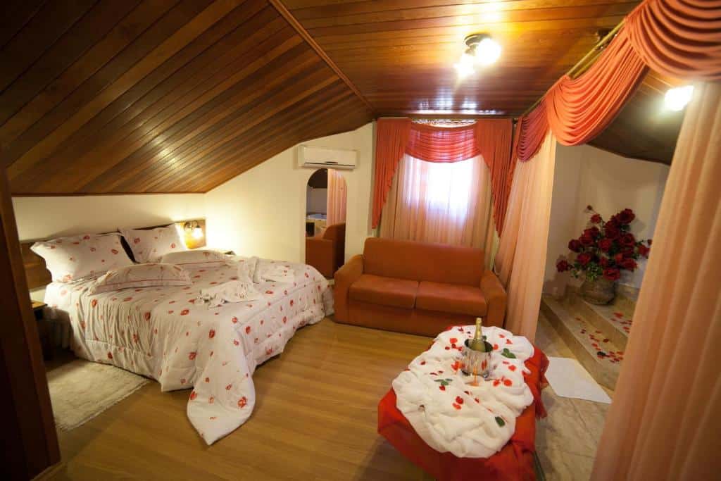 Quarto no sótão com teto de madeira, cama de casal, sofá laranja, mesa com bebidas e cortinas decorativas vermelhas e flores, na Pousada Vollweiter em Entre Rios, para representar pousadas no interior do Paraná.