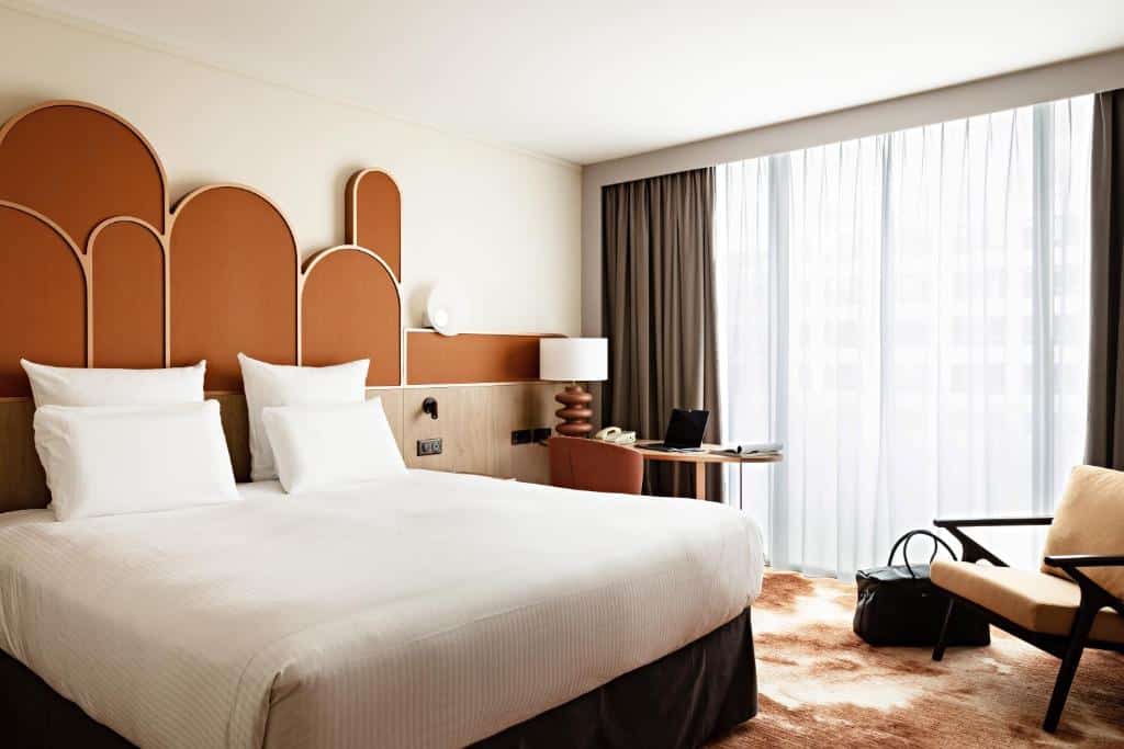 Um quarto no Pullman Paris Centre - Bercy com uma cama grande, duas cadeiras, uma escrivaninha e uma janela com cortinas transparentes que permitem a entrada de luz natural, para representar hotéis com piscina em Paris.